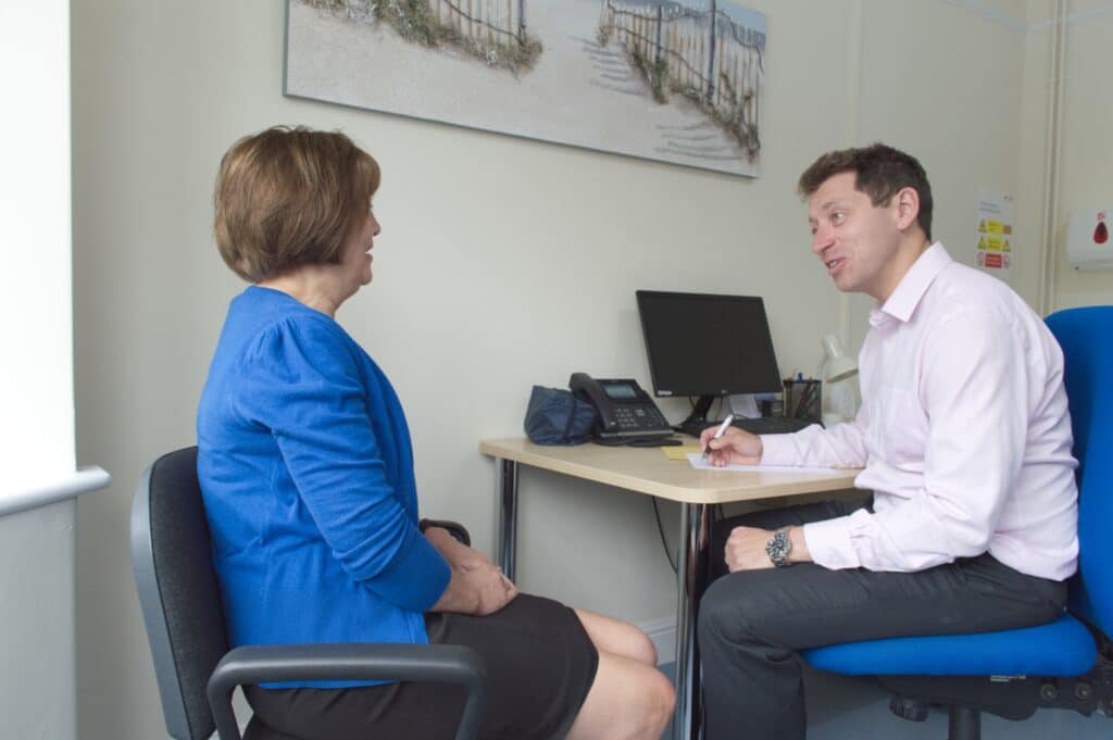 A woman sits opposite a man at a desk in an office, having a conversation. The man is holding a pen and notepad. A computer and telephone are on the desk.