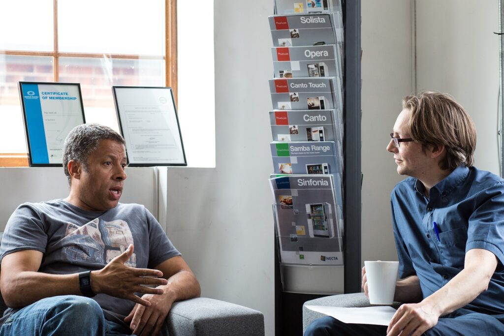 Two men are sitting in chairs having a conversation; one gestures with his hand while the other listens, holding a cup. Brochures and framed certificates are visible in the background.